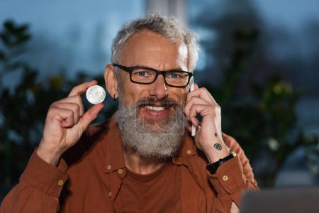 Foto de KYIV, UCRANIA - 17 de noviembre de 2022: feliz hombre de negocios barbudo en gafas que habla en el teléfono inteligente y muestra bitcoin - Imagen libre de derechos