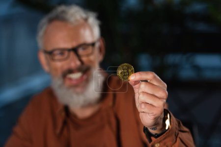 Foto de KYIV, UCRANIA - 17 de noviembre de 2022: alegre hombre de negocios borroso en gafas que muestran bitcoin de oro - Imagen libre de derechos