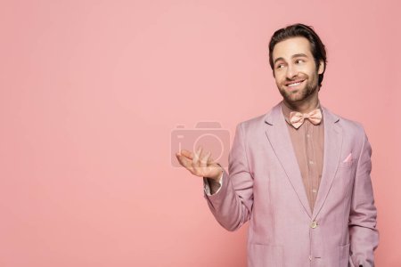 Young host of event in jacket and bow tie pointing with finger isolated on pink 
