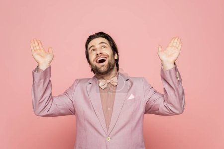 Cheerful host of event in jacket and suit looking up on pink background 