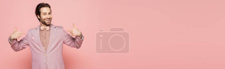 Positive host of event pointing with fingers and looking at camera on pink background, banner 
