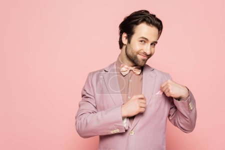 Smiling host of event in jacket touching handkerchief isolated on pink 