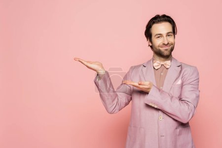Positive Moderatorin in Sakko und Fliege, die mit den Händen auf rosa Hintergrund zeigt 
