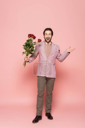 Excited host of event in jacket and bow tie holding bouquet of roses on pink background 