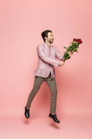Full length of positive host of event holding roses while jumping on pink background 