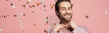 Positive host of event adjusting bow tie near confetti on pink background, banner 