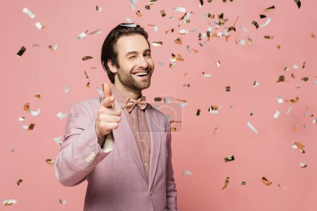 Photo for Smiling host of event pointing at camera under falling confetti on pink background - Royalty Free Image