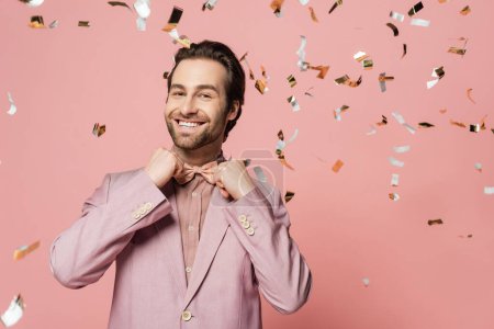 Smiling host of event in jacket adjusting bow tie under confetti on pink background 