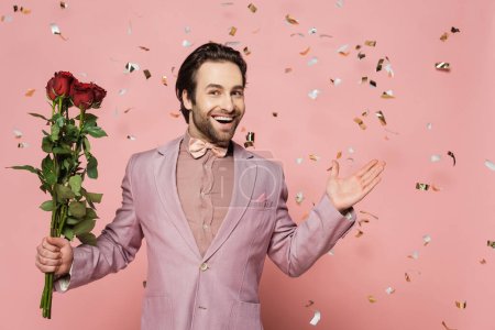 Hôte excité tenant des roses et pointant avec la main sous confettis sur fond rose 