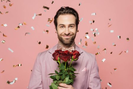 Portrait of positive host of event holding roses under falling confetti on pink background 