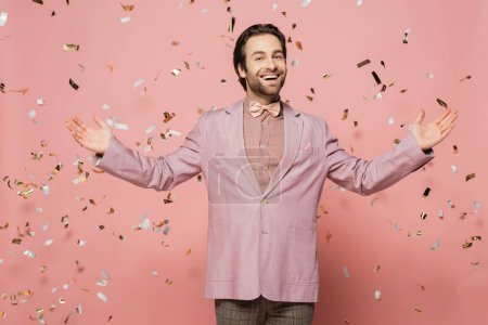 Cheerful host of event pointing at falling confetti and looking at camera on pink background 