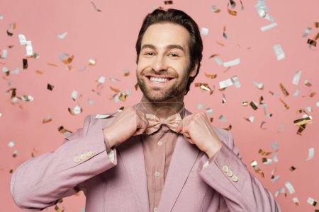 Smiling host of event adjusting bow tie near blurred confetti on pink background 