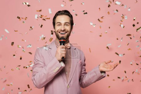 Positive host of event holding microphone under falling confetti on pink background 