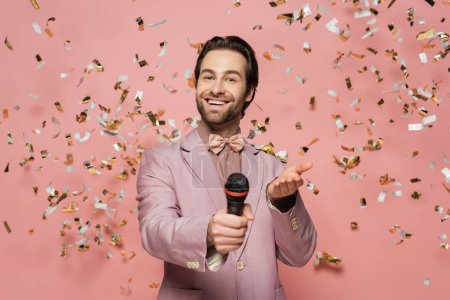 Anfitrión sonriente de evento sosteniendo micrófono y apuntando a la cámara bajo confeti sobre fondo rosa 