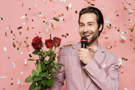 Cheerful host of event talking while holding microphone and roses on pink background with confetti 