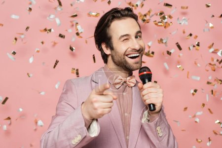 Positive host of event holding microphone and winking at camera under confetti on pink background 