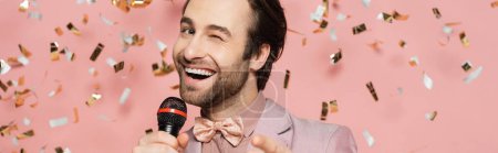Stylish host of event holding microphone and winking at camera under confetti on pink background, banner 