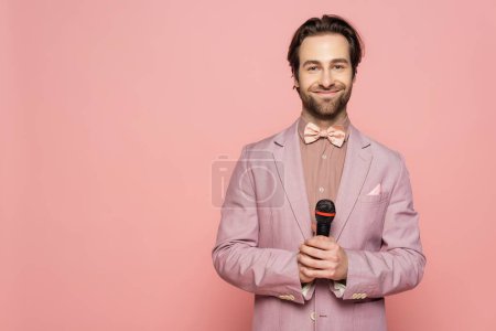 Young host of event holding microphone and looking at camera isolated on pink 