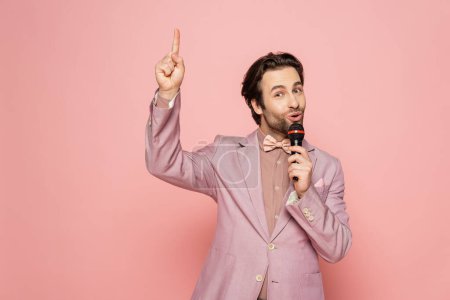 Young and stylish host of event holding microphone while having idea isolated on pink 