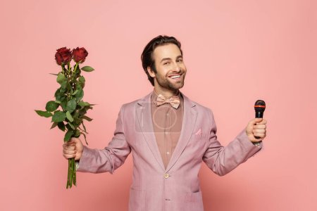 Stylish host of event in jacket and bow tie holding microphone and flowers on pink background 