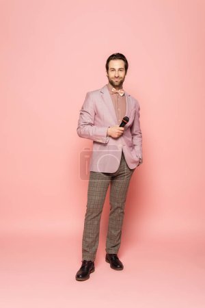 Photo for Full length of trendy host of event holding microphone and looking at camera on pink background - Royalty Free Image