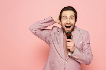 Excited host of event in jacket holding microphone and hand near head isolated on pink 