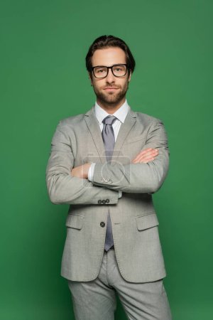 Bearded newscaster in eyeglasses and suit crossing arms on green background 
