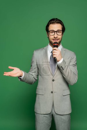 Téléchargez les photos : Ancre de nouvelles en costume parlant au microphone et pointant avec la main sur fond vert - en image libre de droit