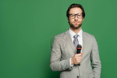 Brunette newscaster in eyeglasses holding microphone and looking at camera on green background 