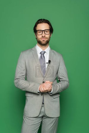 junger Nachrichtensprecher in Anzug und Clip-Mikrofon am Blazer stehend mit geballten Händen auf grün 