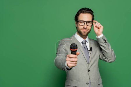 Nachrichtensprecher im Anzug justiert Brille und hält Mikrofon auf grünem Hintergrund 