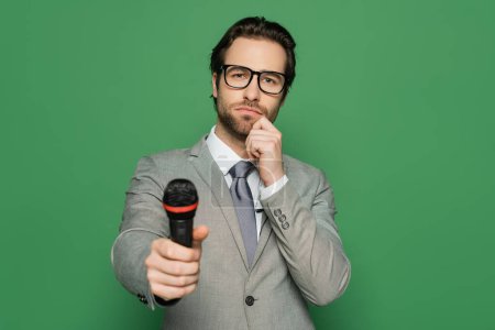 Nachdenklicher Nachrichtensprecher in Anzug und Brille, Mikrofon isoliert auf Grün 
