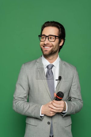 happy news anchor in suit and eyeglasses holding microphone isolated on green 