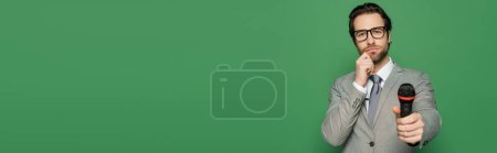 Photo for Pensive news anchor in suit and eyeglasses holding microphone isolated on green, banner - Royalty Free Image