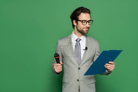diffuseur en costume et lunettes tenant presse-papiers et microphone sur vert 