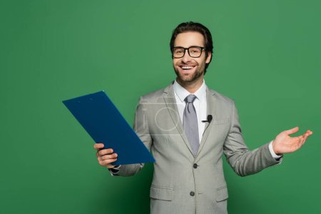 happy news anchor in suit and eyeglasses holding clipboard while gesturing on green background 