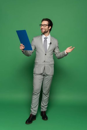 full length of cheerful news anchor in suit and eyeglasses holding clipboard while gesturing on green 