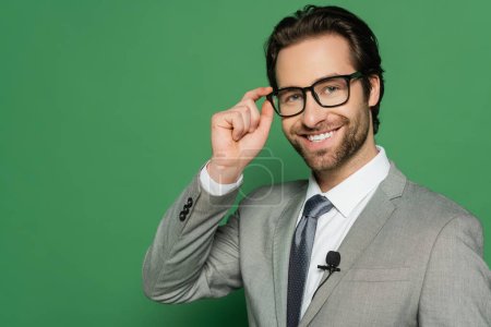 news anchor in suit adjusting eyeglasses while smiling on green background