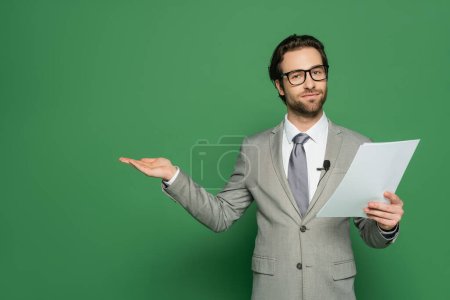 news anchor in eyeglasses and suit holding blank paper and pointing with hand on green