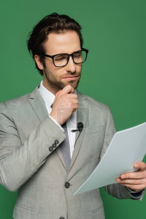 Nachdenklicher Nachrichtenanker in Brille und Anzug mit Blick auf leeres Papier isoliert auf Grün