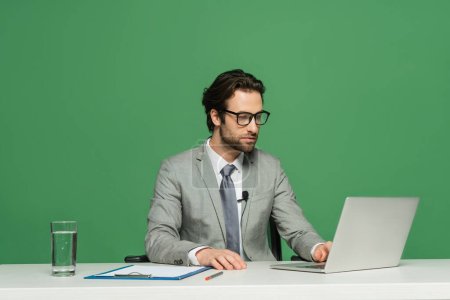 bearded news anchor in eyeglasses and suit using laptop isolated on green