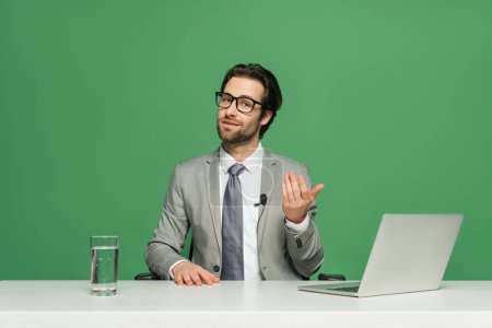 bearded news anchor in eyeglasses and suit talking near laptop isolated on green