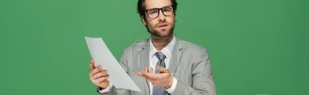 Photo for Confused news anchor in eyeglasses and suit pointing with hand at blank paper isolated on green, banner - Royalty Free Image