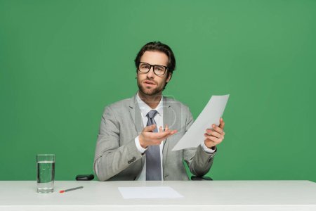 confused news anchor in eyeglasses and suit pointing with hand at blank paper isolated on green