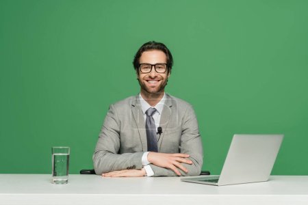 bearded news anchor in eyeglasses and suit smiling while sitting near laptop isolated on green