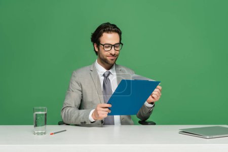 smiling news anchor in eyeglasses and suit holding clipboard near laptop isolated on green