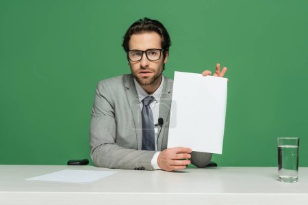 bearded news anchor in eyeglasses and suit holding blank paper isolated on green