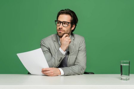 thoughtful broadcaster in eyeglasses and suit holding blank paper isolated on green