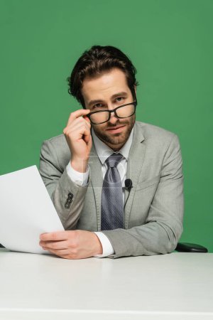 broadcaster in suit adjusting eyeglasses and holding blank paper isolated on green