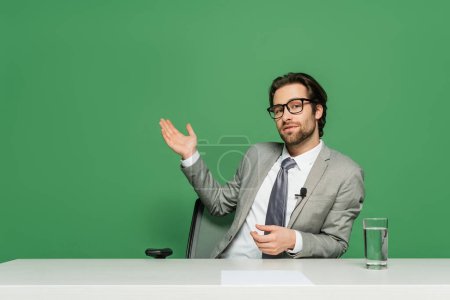 broadcaster in eyeglasses and grey suit sitting at desk and pointing with hand isolated on green